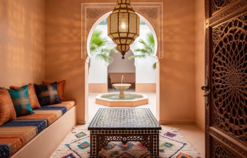 Eye-level photo of a Moroccan riad interior with a hand-pierced brass lantern illuminating tadelakt walls, a Fes-style zellige mosaic table, a carved cedar mashrabiya screen, and colorful Berber textiles; an arched courtyard with palm fronds and a small fountain appears beyond.